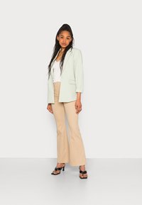 Blazer verde menta, blusa branca, calças largas beges e sandálias de salto alto pretas. O blazer tem mangas longas e um corte solto.