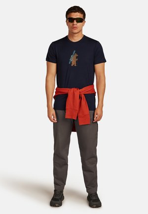 Hombre con gafas de sol oscuras, camiseta azul marino con un gráfico de un oso llevando esquís, pantalones grises, zapatos negros y un sudadera roja atada alrededor de la cintura.