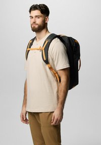 Schwarzer Rucksack mit orangefarbenen Akzenten, ergonomischen Gurten und einem Brustgurt, getragen von einem Mann in einem beigen Hemd und braunen Hosen.