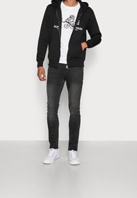 Svart zip-up hoodie med långa ärmar, en framsidan grafisk bild av en dinosaurie, i kombination med slim-fit mörka jeans och vita sneakers.