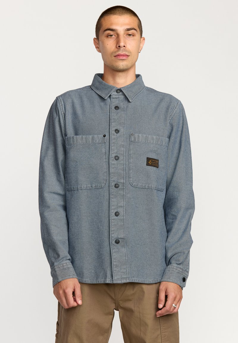 Camisa de manga larga con botones en denim azul claro, con dos bolsillos en el pecho, botones negros y una etiqueta texturizada marrón en el bolsillo izquierdo.