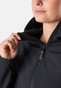 Zwarte softshell jack met een rits kraag, voorzien van een fleece-gevoerd interieur en een hand die de kraag vasthoudt. Soepele textuur en aangesloten ontwerp.