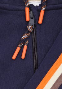 Navyblauwe hoodie met een volledige rits, contrasterende oranje trekkoordtips en horizontale strepen in oranje, crème en bruin.