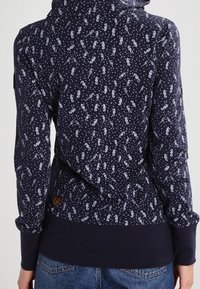 Mörkblå hoodie med vitt sjöhäst- och bubbelmönster, bärs av en person som är vänd ryggen mot kameran, ihop med blå jeans.