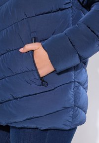 Tummansininen puffatakki, jossa on tilkkutekstuuri. Eteisessä on sivuvetoketjullinen tasku ja elastiset hihansuut. Materiaali näyttää kevyeltä ja kiiltävältä.