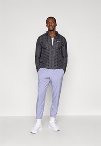 Black synthetic jacket with quilted texture, paired with light purple tapered pants. Both garments feature Nike branding. White sneakers complete the look.