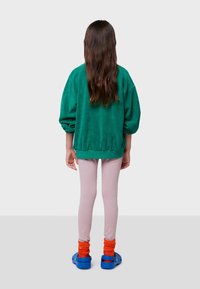 Niño con cabello largo castaño, vistiendo un suéter verde grande, leggings rosa claros, calcetines naranjas y sandalias azules, de pie de espaldas.