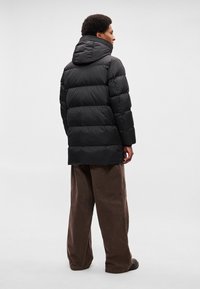 Casaco acolchoado preto com capuz, com secções acolchoadas. O modelo usa calças largas castanhas. O fundo é plano e claro.