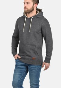 Hematietgrijze hoodie met een buidelzak, beige trekkoorden en een bruin leren patch detail aan de zoom. Zachte, gestructureerde stof.
