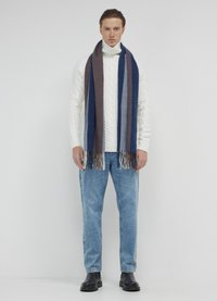 Wollmischung-Schal mit dunkelblauen und braunen diagonalen Streifen sowie Fransen. Das Modell trägt einen dicken weißen Zopfpullover und blaue Jeans.