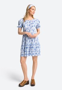 Rochie florală albastră cu mâneci bufante, croială lejeră și tiv în straturi. Asortată cu saboți maroi cu accente metalice.