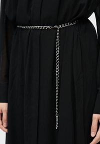 Ασημένια ζώνη από αλυσίδα με πλεγμένο σχέδιο, με μακριά αλυσίδα που κρέμεται στη μία πλευρά, φορεμένη πάνω από μαύρο ένδυμα.