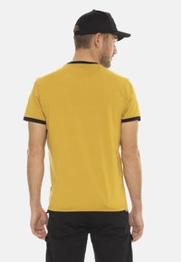 T-shirt jaune à manches courtes avec des accents noirs sur le col et les manches, fabriqué dans un tissu lisse, de style ajusté, avec des détails de couture visibles.