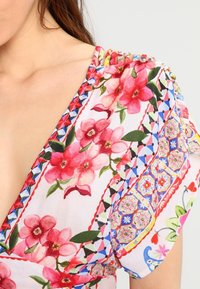 Bluse med blomstermønster i blødt stof. Har pink blomster, grønne blade og farverige geometriske accenter langs Halsudskæringen og ærmerne.