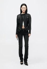 Noor naine seisab seljas must nahkjakk, mustad kõrge vöökohaga teksad, valge lühike topp ja mustad terava ninaga kingad.