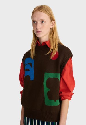 Jeune personne aux longs cheveux blonds porte un pull sans manches marron avec des formes bleues, vertes et rouges par-dessus une chemise rouge à col et un pantalon rayé.