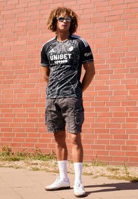 Svart sportski trøje med hvite og grå detaljer, som har "UNIBET" logo, kombinert med mørke cargo shorts, hvite sokker og sneakers.