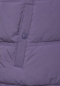Lila puffjaktssektion med en strukturerad, vadderad yta, en horisontell söm och en cirkulär knapp med präglad logotyp.