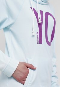 Sudadera de color azul claro con un gran gráfico morado que dice "YO" y el logo de Reebok. Fabricada con un material suave, cuenta con un bolsillo tipo canguro y cordones.