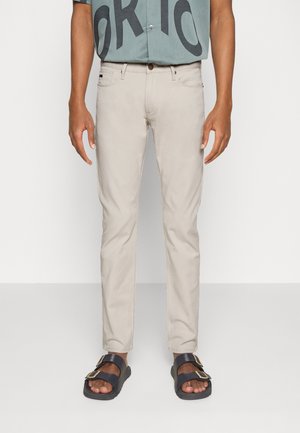 Pantaloni beige realizzati in tessuto leggero con vestibilità slim, abbinati a sandali neri con due cinturini regolabili e una suola strutturata.