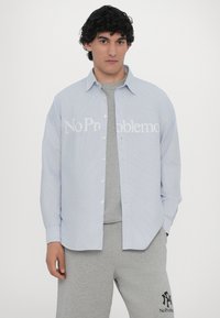 Cămașă cu dungi albastre deschise, cu butoni și textul alb "No Problemo", purtată peste un tricou gri și asortată cu pantaloni de trening gri.