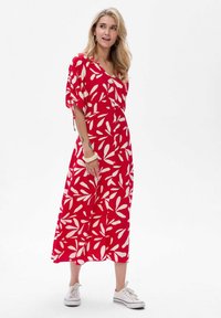 Frau mit langen blonden Haaren, die ein rotes Maxikleid mit weißen Blattmustern, weiße Turnschuhe und Ohrreifen trägt, vor einem weißen Hintergrund stehend.