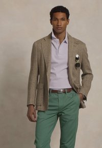 Brun herringbone-blazer over en lys lilla polo, kombinert med grønne bukser. Tilbehøret inkluderer solbriller og en sølv beltespenne.