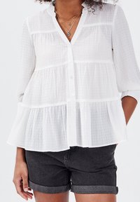 Blouse blanche semi-transparente en tissu texturé et froissé, avec un ourlet à volants, des manches longues et une fermeture boutonnée à l'avant. Associée à un short gris.