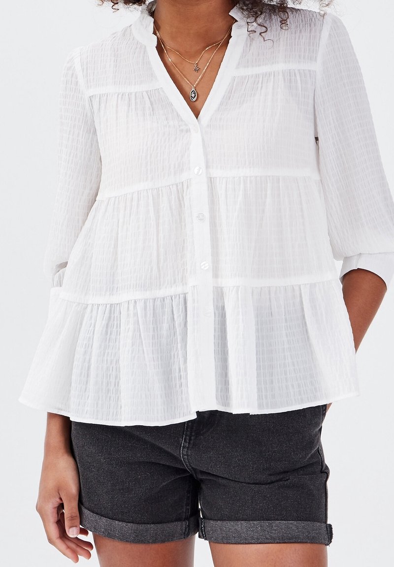 Blouse blanche semi-transparente en tissu texturé et froissé, avec un ourlet à volants, des manches longues et une fermeture boutonnée à l'avant. Associée à un short gris.