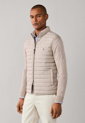Homme portant un gilet matelassé beige sans manches sur un pull beige clair et un pantalon blanc, debout devant un fond uni gris clair.
