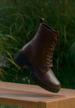 Brauner Leder-Schnürstiefel mit dicker schwarzer Sohle, der über einer Holzoberfläche mit grüner Vegetation im Hintergrund schwebt.
