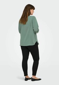 Minta zaļa blūze ar garām piedurknēm, noapaļots apakšmalas dizains, nēsāta kopā ar melnajām šaurajām biksēm un melnajām plakanajām kurpēm. Audums izskatās gluds un viegls.