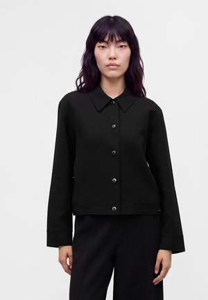 Femme aux longs cheveux noirs portant une veste noire boutonnée et un pantalon noir, debout devant un fond clair uni.