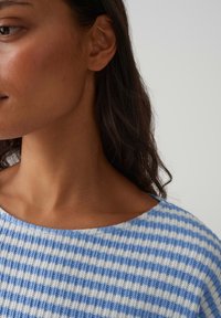 Maglione a quadretti blu e bianco con scollo rotondo, trama a coste e tessuto morbido. Inquadratura sul collo superiore e sul volto in un ambiente neutro.