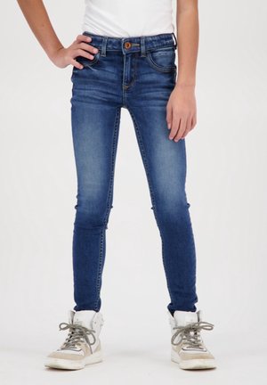 Ciemnoniebieskie slim-fit dżinsy z zapięciem na guzik, pięcioma kieszeniami i lekkim wyblaknięciem. Stylizowane z białą koszulą i szarymi sneakersami typu high-top.