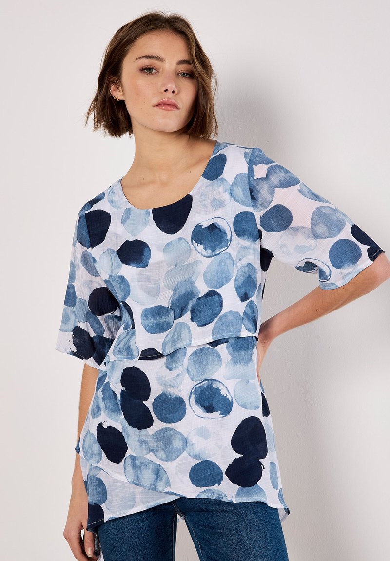 Blue and white patterned top featuring large abstract circles, short sleeves, and a scoop neckline, made from lightweight fabric.