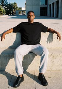 Junger Mann in schwarzem T-Shirt und grauer Jogginghose sitzt auf einer Betonbank im Freien mit den Armen auf der Lehne in sonniger urbaner Umgebung.