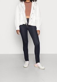 Chaqueta blanca recortada con botones en la parte frontal y dos bolsillos, combinada con una blusa de rayas marrón y naranja y jeans oscuros ajustados. Zapatillas blancas con acentos en tonos pastel.