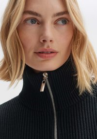 Blonde Frau trägt einen schwarzen Rippstrick-Turtleneck-Pullover mit silbernem Reißverschluss und schaut leicht zur Seite vor einem schlichten weißen Hintergrund.