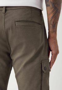 Pantalons cargo en tissu vert olive avec une texture lisse. Comprend une poche arrière et une poche latérale à rabat avec un détail de bouton noir.