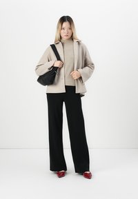 Woman wearing beige coat and sweater, black wide-leg pants, red shoes, and carrying a black shoulder bag against white background.