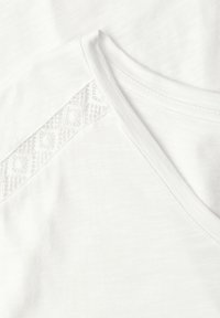 Chemise blanche à col en V avec un délicat détail en dentelle le long de la couture de l'épaule, montrant une texture de tissu douce et une couture fine.