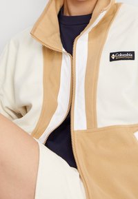 Fleecejacka i krämfärgad och tan, med hög krage, dragkedjestängning och Columbia-logotyp. Mjuk textur med kontrasterande vita paneler.