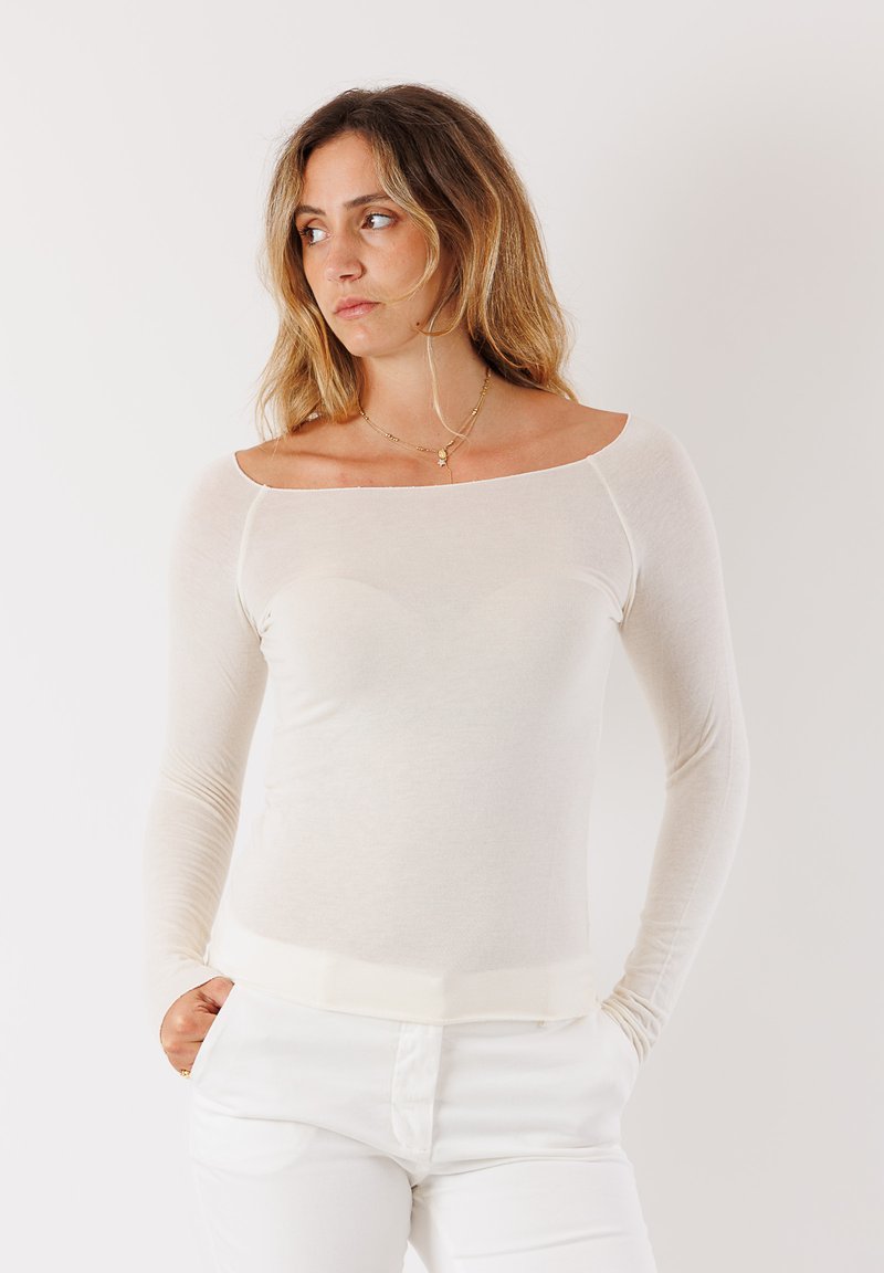 Top lungo a maniche lunghe color beige chiaro con scollatura a spalle scoperte, realizzato in un tessuto morbido e leggero. Il design aderente esalta la forma del corpo.