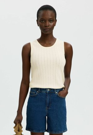 Femme debout face à l'objectif, portant un haut en maille crème sans manches et un short en jean, tenant un petit sac texturé, une main dans la poche, fond neutre.