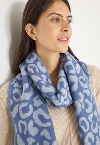 Écharpe bleue avec un motif léopard bleu clair, en matière douce, élégamment drapée autour du cou sur un pull beige.