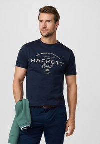 Hombre con cabello corto y castaño, vistiendo una camiseta azul marino "Hackett Sport", sosteniendo una sudadera verde doblada, de pie frente a un fondo claro y liso.
