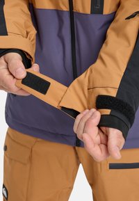 Skijacke aus braunem und lila Stoff, mit schwarzen Akzenten, verstellbaren Bündchen mit Klettverschluss und durchgehendem Reißverschluss. Zeigt ein strukturiertes Material.