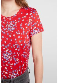 Camiseta roja con flores y mangas cortas. Presenta pequeños patrones de flores blancas y azules. Tejido suave y cuello redondo, combinada con pantalones grises.