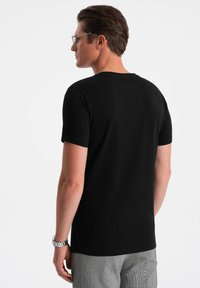 Sort kortærmet t-shirt med rund halsudskæring, lavet af glat stof. Bæres med grå mønstrede bukser og har et ur på håndleddet.